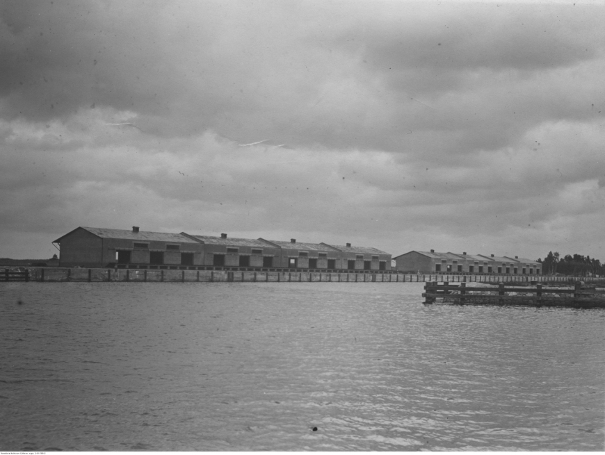 Zdjęcie basenu amunicyjnego na Westerplatte wykonane od strony wejścia kanału portowego. Wyraźnie widoczne całe założenie wraz z magazynami stojącymi wokół. Obiekty na zdjęciu są w tzw. stanie surowym, bez drzwi i okien, które dopiero będą montowane. Zdjęcie wykonano w drugiej połowie 1926 r. (Narodowe Archiwum Cyfrowe, NAC)