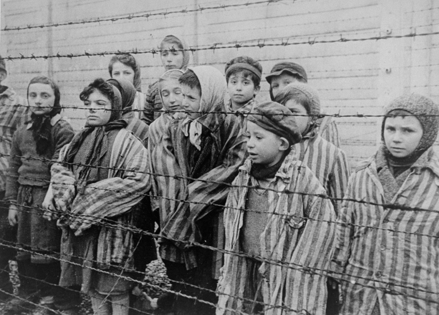 Ocalałe z Auschwitz dzieci, ubrane w obozowe kurtki dla dorosłych, stoją za ogrodzeniem z drutu kolczastego (Źródło: Domena publiczna)